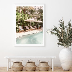May include: A framed print of a pool scene with striped umbrellas, lounge chairs, and lush greenery. The pool has clear, turquoise water. The print is displayed on a white wall above a bench with three woven baskets.
