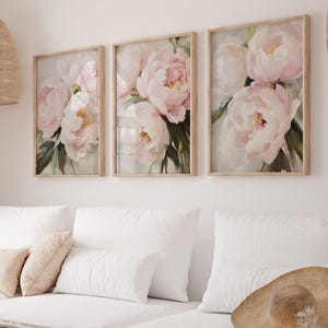 May include: Three framed floral art prints featuring soft pink peonies with green leaves. The artwork is displayed above a white sofa with pillows. The frames are light-colored wood.