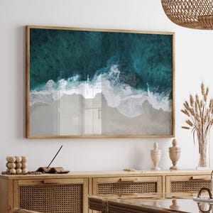 May include: A framed print depicting an aerial view of a beach. The artwork shows turquoise ocean waves breaking onto a sandy shore. The print is enclosed in a light brown wooden frame, adding a natural touch to the coastal scene.