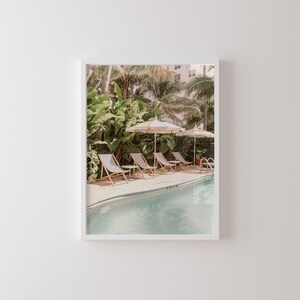 May include: A framed print featuring a serene poolside scene. The image showcases striped lounge chairs, cream-colored umbrellas, and a turquoise pool surrounded by lush tropical foliage. A building is visible in the background, adding depth to the composition.