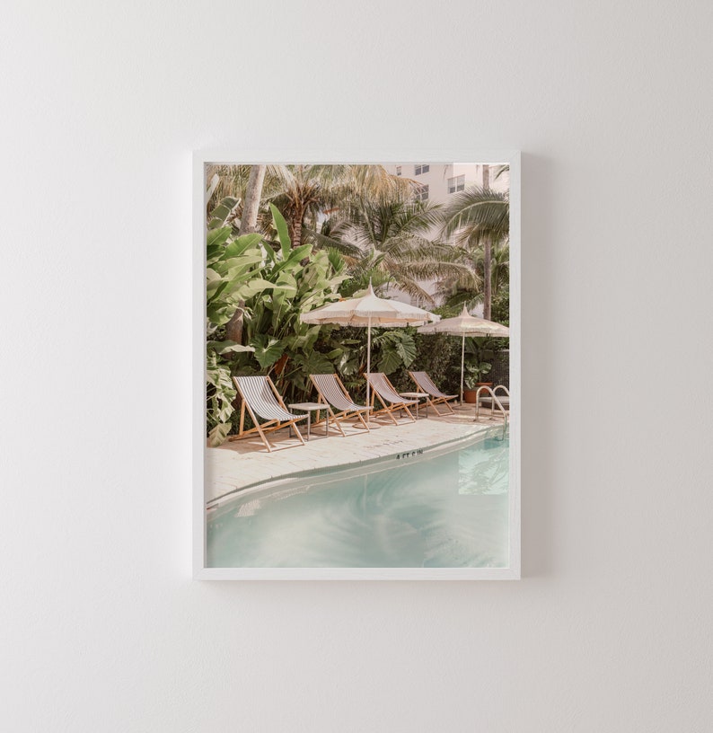 May include: A framed print featuring a serene poolside scene. The image showcases striped lounge chairs, cream-colored umbrellas, and a turquoise pool surrounded by lush tropical foliage. A building is visible in the background, adding depth to the composition.