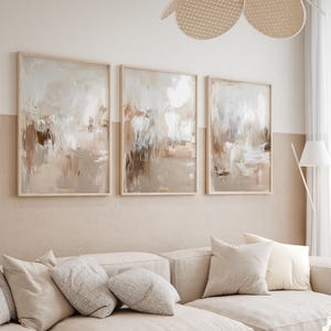 May include: Three abstract paintings in beige, brown, and white tones are displayed in wooden frames on a light beige wall. The paintings are arranged in a row, with the leftmost painting being slightly larger than the other two.