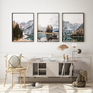 May include: Three framed landscape photographs of a lake surrounded by mountains and trees. The photos are hung on a white wall above a wooden cabinet with shelves.