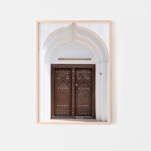 May include: A framed photograph of a brown wooden door with intricate carvings and a metal handle. The door is set within a white arched doorway.