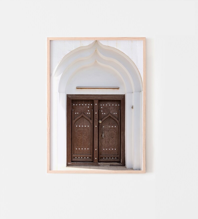 May include: A framed photograph of a brown wooden door with intricate carvings and a metal handle. The door is set within a white arched doorway.