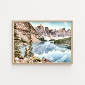 May include: A framed landscape print featuring a serene lake reflecting towering, snow-capped mountains. The image showcases a vibrant blue sky, lush green trees, and the natural beauty of the wilderness. The frame is a light brown color.