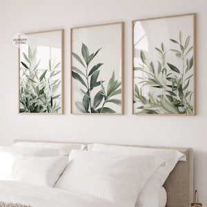 May include: Three framed botanical prints featuring green and gray leaves. The prints are in light brown wooden frames. The artwork is displayed above a bed with white bedding. The prints are from the "JAMHOME STUDIO".