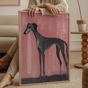 Puede incluir: Obra de arte enmarcada que representa un galgo en tonos grises sobre un fondo rosa. El perro est&aacute; pintado en un estilo minimalista con patas largas y delgadas. La obra de arte est&aacute; encerrada en un marco de madera de color claro.