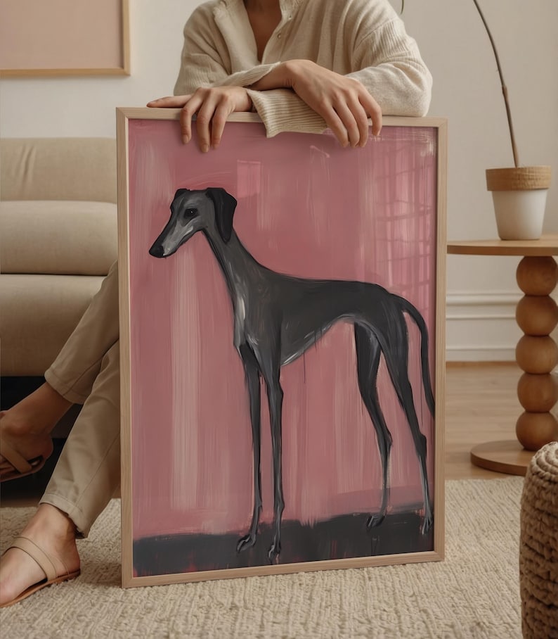 Puede incluir: Obra de arte enmarcada que representa un galgo en tonos grises sobre un fondo rosa. El perro est&aacute; pintado en un estilo minimalista con patas largas y delgadas. La obra de arte est&aacute; encerrada en un marco de madera de color claro.