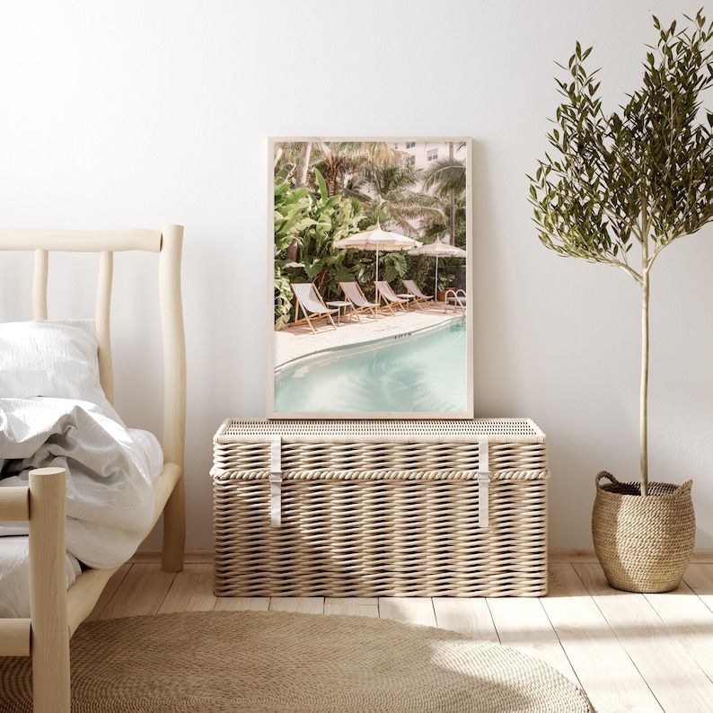 May include: A framed print depicts a serene pool scene with lounge chairs and striped umbrellas. Lush greenery surrounds the pool, creating a tropical ambiance. The print rests on a woven trunk, with a bed frame and a potted plant in the foreground.