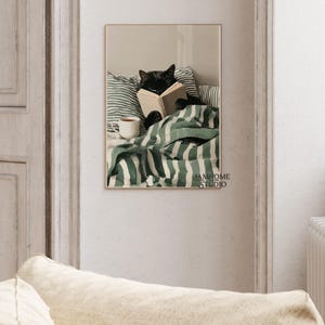 May include: A framed print featuring a black cat reading a book in bed. The cat is nestled under a green and white striped blanket, with a matching pillow behind it. A white mug of tea sits nearby. The print has a gold frame.