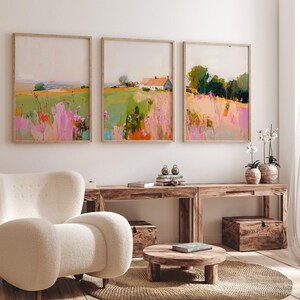 May include: Three framed landscape paintings displayed in a row, featuring abstract fields with pink, green, and orange brushstrokes. The center painting depicts a small white house with a red roof nestled within the colorful meadow scenery.