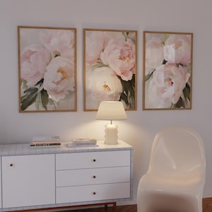 May include: Three framed floral art prints featuring soft pink peonies with green leaves. The artwork is displayed above a white cabinet with a lamp and a modern white chair. The frames have a light brown wood finish.