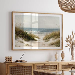 Puede incluir: Una impresión enmarcada de un paisaje de playa con un camino que conduce al océano. La obra presenta dunas de arena con hierbas altas en tonos verdes y beige, y un cielo y un océano azul suave.