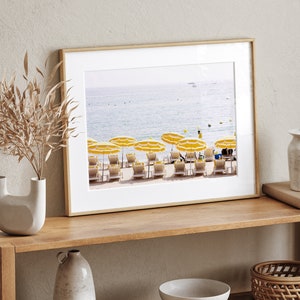 Puede incluir: Impresi&oacute;n enmarcada de una escena de playa con sombrillas a rayas amarillas y blancas y tumbonas. El oc&eacute;ano es visible en el fondo, con un cielo azul claro. La impresi&oacute;n est&aacute; en un marco de madera, exhibida en un estante de madera.