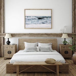 Op de afbeelding: Een houten bedframe met een wit dekbed en kussens. Het bed heeft een houten hoofdbord en voetbord. Boven het bed hangt een ingelijst foto van oceaan golven. Twee houten nachtkastjes met lampen staan aan weerszijden van het bed.