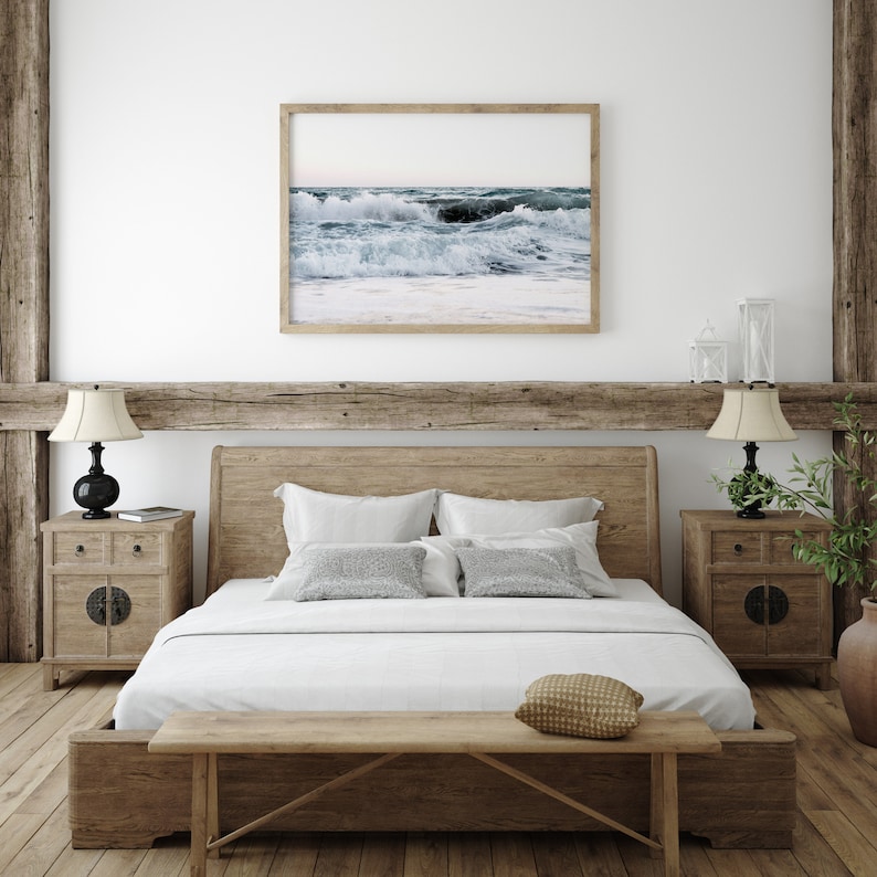Op de afbeelding: Een houten bedframe met een wit dekbed en kussens. Het bed heeft een houten hoofdbord en voetbord. Boven het bed hangt een ingelijst foto van oceaan golven. Twee houten nachtkastjes met lampen staan aan weerszijden van het bed.