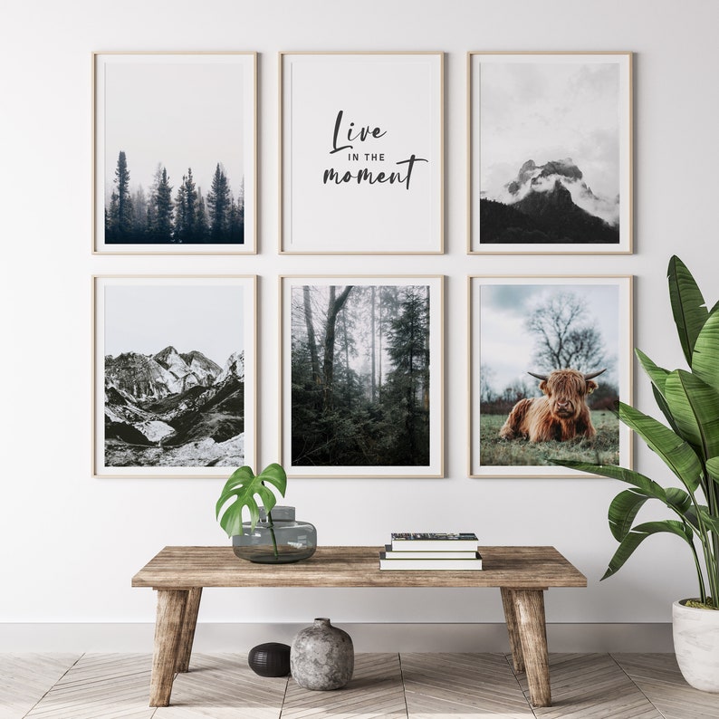 May include: A set of six framed prints featuring nature scenes. The prints include a mountain range, a forest, a cloudy mountain peak, a forest with fog, a highland cow, and a text print that reads "Live in the moment".