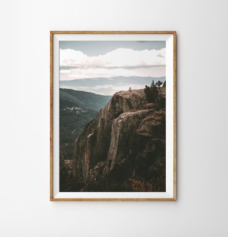 May include: A framed photograph of a rocky cliff face with a view of a forest and distant mountains. The sky is cloudy and the colors are muted.