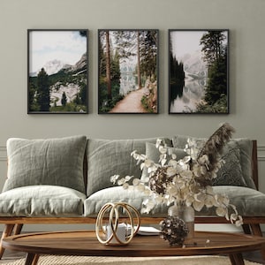May include: Three framed landscape photographs of a lake surrounded by trees and mountains. The photos are hung on a light green wall above a light brown couch with three pillows.