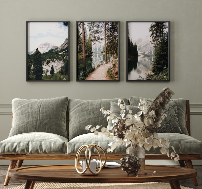 May include: Three framed landscape photographs of a lake surrounded by trees and mountains. The photos are hung on a light green wall above a light brown couch with three pillows.