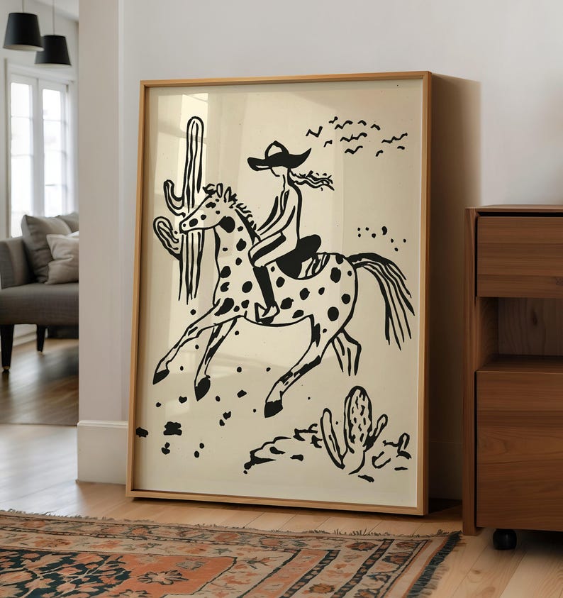 May include: Black and white illustration of a person riding a horse with a cactus in the background. The image is framed in a light wood frame.