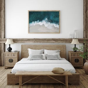 May include: A bedroom scene featuring a wooden bed frame, nightstands, and a bench. A framed aerial view of a beach with turquoise water and white sand hangs above the bed. The room has a neutral color palette with natural wood accents.