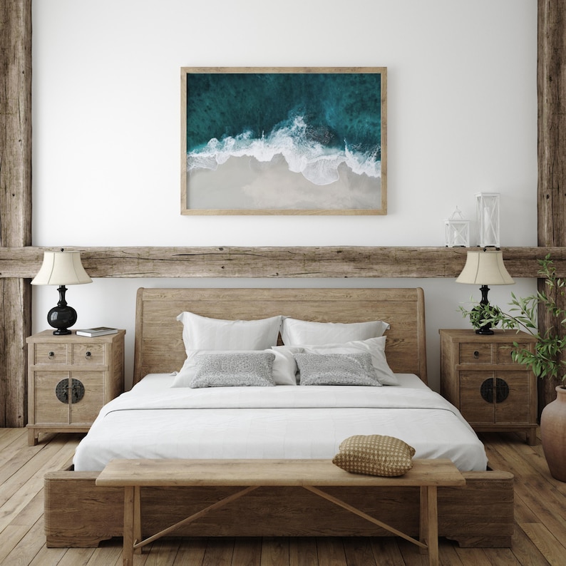 May include: A bedroom scene featuring a wooden bed frame, nightstands, and a bench. A framed aerial view of a beach with turquoise water and white sand hangs above the bed. The room has a neutral color palette with natural wood accents.