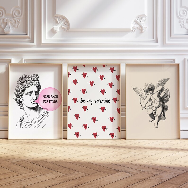 May include: Three framed prints with Valentine's Day themes. The left print features a black and white illustration of a man blowing a bubble that says "More Amor Por Favor". The center print has a white background with a repeating pattern of red hearts with arrows. The text "be my valentine" is in the center of the print. The right print features a black and white illustration of a cherub with wings and an arrow.