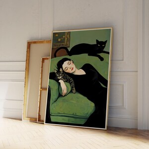 May include: A framed art print depicts a woman in a black dress reclining on a green sofa with a tabby cat. A black cat rests on the sofa's back. The artwork has a vintage aesthetic, with a gold frame and a neutral background.