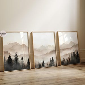 May include: Three vertical framed wall art prints featuring a watercolor-style landscape of misty mountains and dark evergreen pine trees in shades of beige, gray, and black. A small circular logo with the text AMHOME STUDIO is visible.
