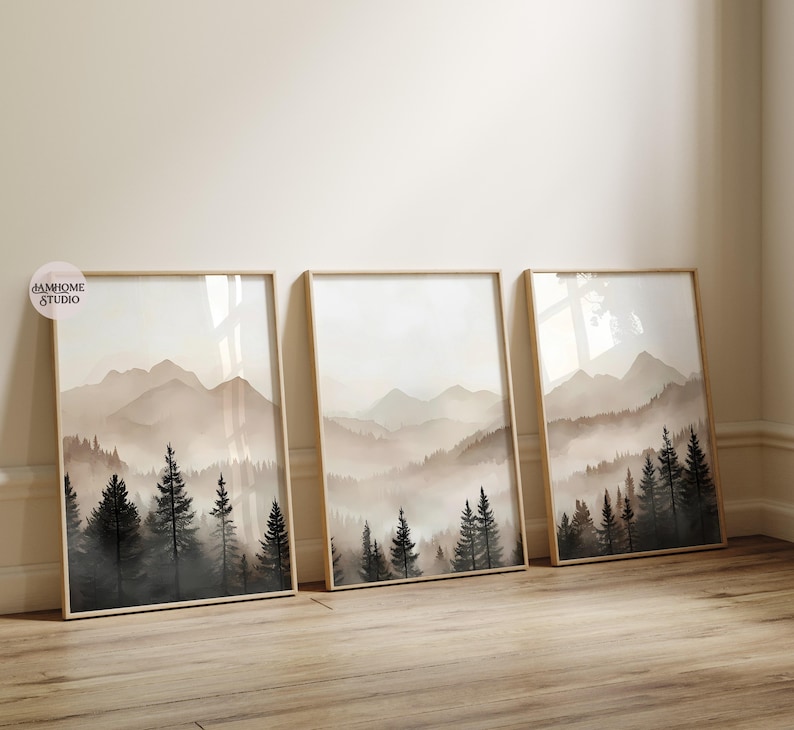 May include: Three vertical framed wall art prints featuring a watercolor-style landscape of misty mountains and dark evergreen pine trees in shades of beige, gray, and black. A small circular logo with the text AMHOME STUDIO is visible.