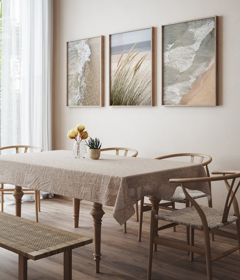 May include: A dining room with a light brown wooden table and chairs, a light brown wooden bench, and a beige tablecloth. Three framed prints of beach scenes hang on the wall behind the table.