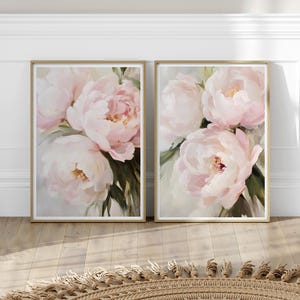 以下が含まれることがあります： 2つの額入りアートプリント。柔らかなピンク色の牡丹と緑の葉が描かれています。絵画は金色のフレームに収められ、明るい背景に配置されています。アートワークは明るい木製の床に展示されています。