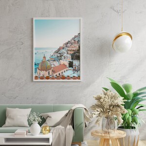 May include: A framed photograph of a colorful Italian village built on a hillside overlooking the ocean. The photo is hanging on a white wall above a green sofa with a white throw blanket.