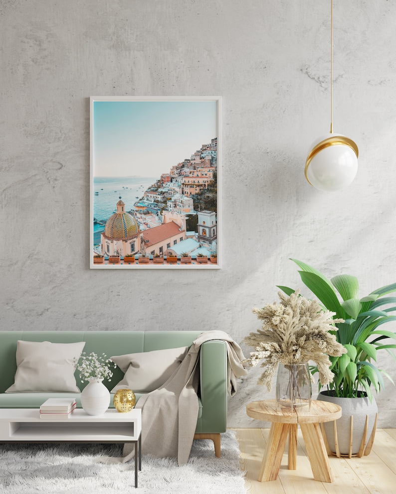 May include: A framed photograph of a colorful Italian village built on a hillside overlooking the ocean. The photo is hanging on a white wall above a green sofa with a white throw blanket.