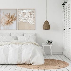 May include: A white bed with a white duvet cover and two white pillows. There are two framed prints on the wall above the bed, one with a beach scene and the other with a bunch of dried grass. A white bedside table with a potted plant and a book sits next to the bed. A woven rug is on the floor in front of the bed. A white cabinet with louvered doors is in the background.