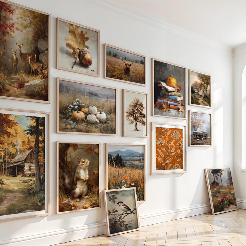May include: A collection of framed art prints displayed on a white wall. The prints feature various autumnal scenes, including landscapes, still lifes with pumpkins, and wildlife like deer and squirrels. The frames are a light wood color.