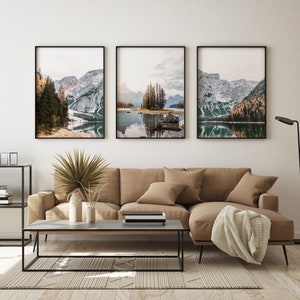 May include: Three framed landscape photographs of a lake surrounded by snow-capped mountains and trees. The photos are hung on a white wall above a brown couch and a coffee table.