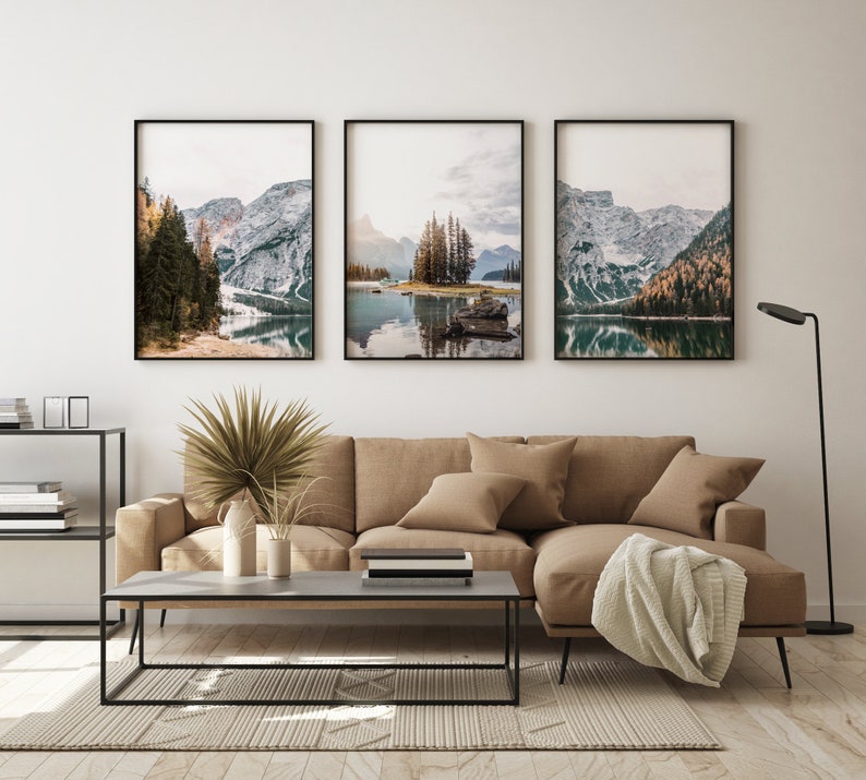 May include: Three framed landscape photographs of a lake surrounded by snow-capped mountains and trees. The photos are hung on a white wall above a brown couch and a coffee table.