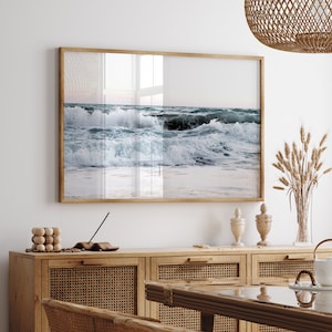 Op de afbeelding: Een ingelijste foto van oceaan golven die breken op een zandstrand. De foto is in een licht houten lijst en hangt aan een witte muur boven een houten kast.