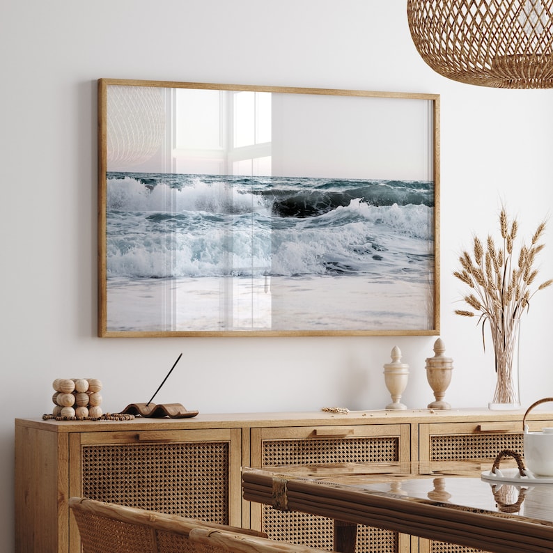 Op de afbeelding: Een ingelijste foto van oceaan golven die breken op een zandstrand. De foto is in een licht houten lijst en hangt aan een witte muur boven een houten kast.