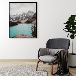 May include: A framed photograph of a mountain lake with snow-capped peaks in the background. The lake is a vibrant turquoise blue and the mountains are a mix of brown and white. The photo is hanging on a white wall above a gray armchair with a brown throw blanket.