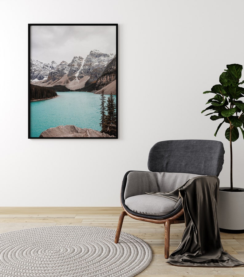 May include: A framed photograph of a mountain lake with snow-capped peaks in the background. The lake is a vibrant turquoise blue and the mountains are a mix of brown and white. The photo is hanging on a white wall above a gray armchair with a brown throw blanket.
