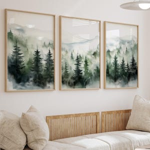 May include: Three framed watercolour paintings of a misty forest scene with green trees and a light blue sky. The paintings are in wooden frames and are hung on a white wall.