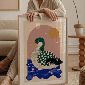 May include: Framed art print with a duck illustration. The duck has a green head and wings with pink polka dots, a yellow beak, and is set against a pink and beige background with a yellow sun and blue water. The print is in a wooden frame.