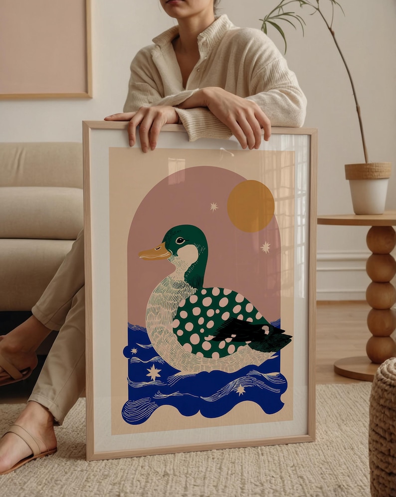 May include: Framed art print with a duck illustration. The duck has a green head and wings with pink polka dots, a yellow beak, and is set against a pink and beige background with a yellow sun and blue water. The print is in a wooden frame.