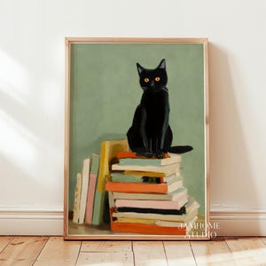 Puede incluir: Una impresión artística enmarcada que muestra un gato negro sentado sobre una pila de libros coloridos. El gato tiene ojos amarillos y está sobre un fondo verde apagado. La obra de arte está en un marco de madera y se exhibe sobre un suelo de madera.