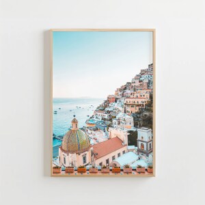 May include: A colorful cityscape with a white domed church building on a cliff overlooking the ocean. The buildings are painted in shades of pink, yellow, and blue. The ocean is a light blue color and there are boats in the distance.