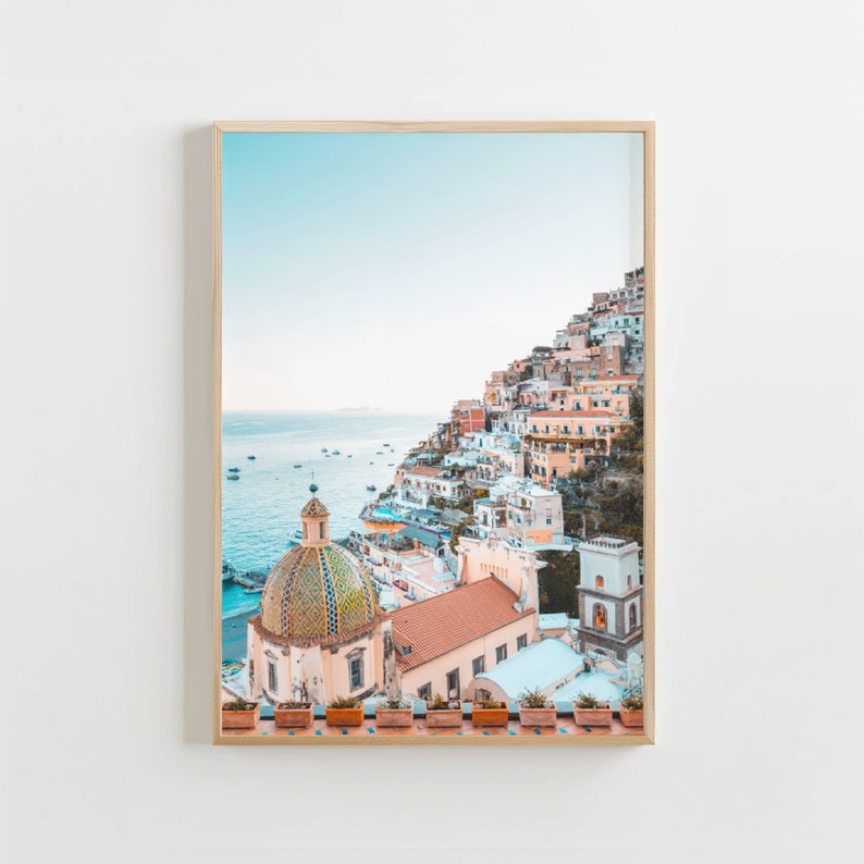 May include: A colorful cityscape with a white domed church building on a cliff overlooking the ocean. The buildings are painted in shades of pink, yellow, and blue. The ocean is a light blue color and there are boats in the distance.